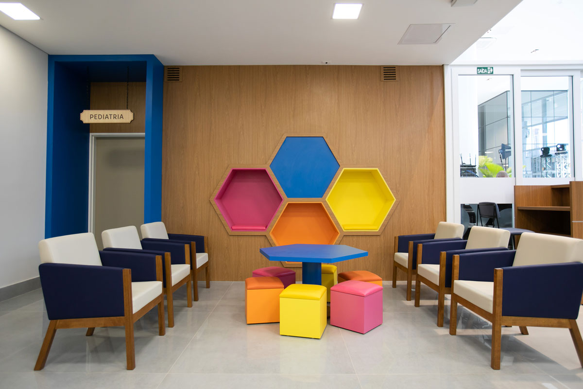 Sala de recepção pediátrica, onde se vê poltronas nas laterais e ao centro móveis coloridos.