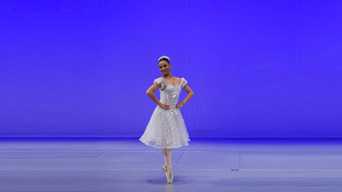 aluna Julia está ao centro da imagem, com figurino branco de ballet, ela está com as mãos na cintura e nas pontas dos pés. O fundo é azul e o chão é cinza claro.